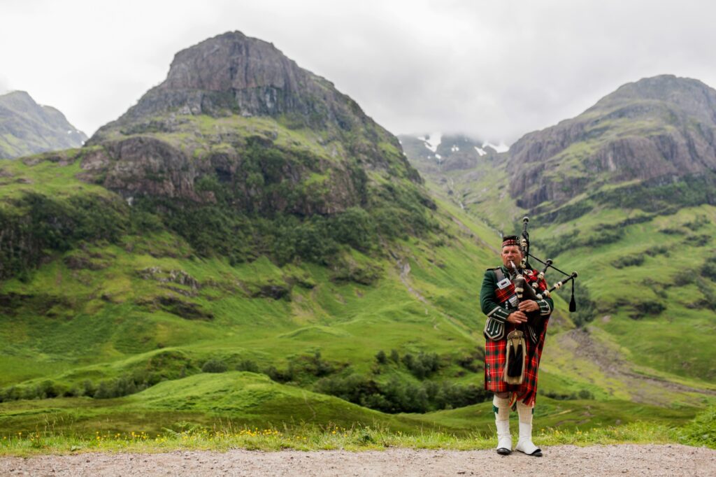 A Scottish bagpiper in traditional Highland dress plays the bagpipes before the mountains of Glen Coe, Scotland